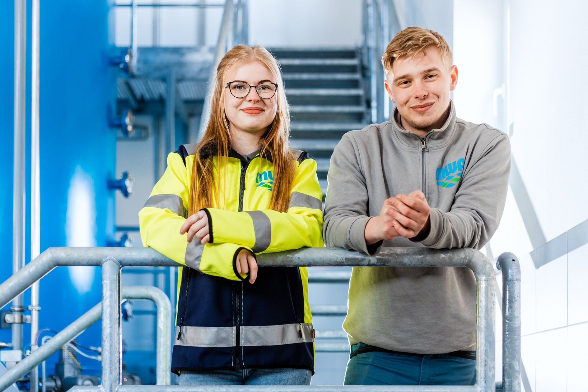 Zwei junge Leute (männlich und weiblich) stehen in einem Wasserwerk an einer Brüstung und schauen in die Kamera.