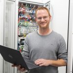 Mann mit Laptop in der Hand vor Schaltschrank. Lächelt in Kamera.