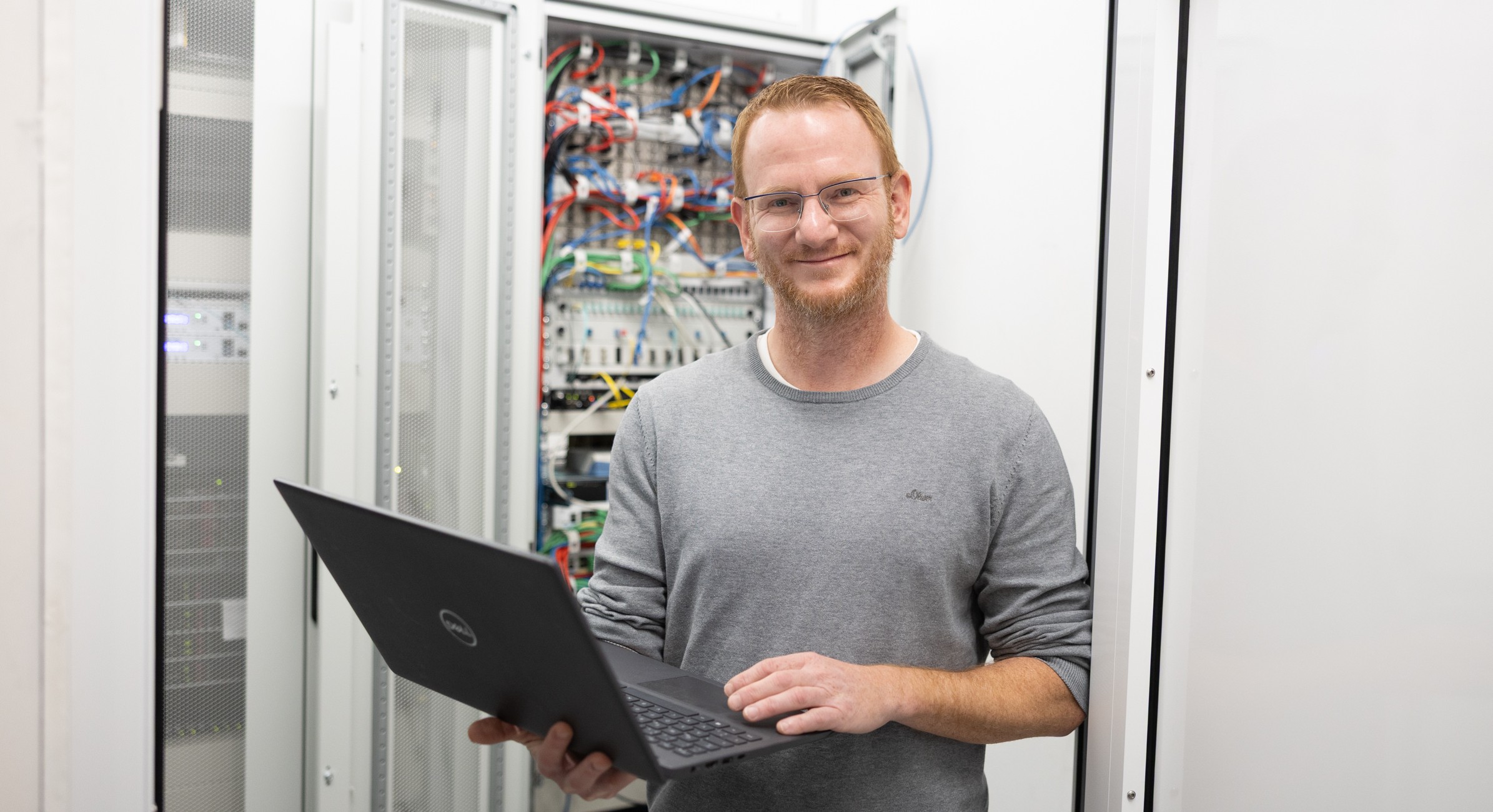 Mann mit Laptop in der Hand vor Schaltschrank. Lächelt in Kamera.