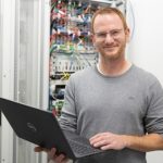 Mann mit Laptop in der Hand vor Schaltschrank. Lächelt in Kamera.