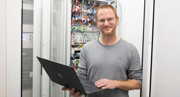Mann mit Laptop in der Hand vor Schaltschrank. Lächelt in Kamera.