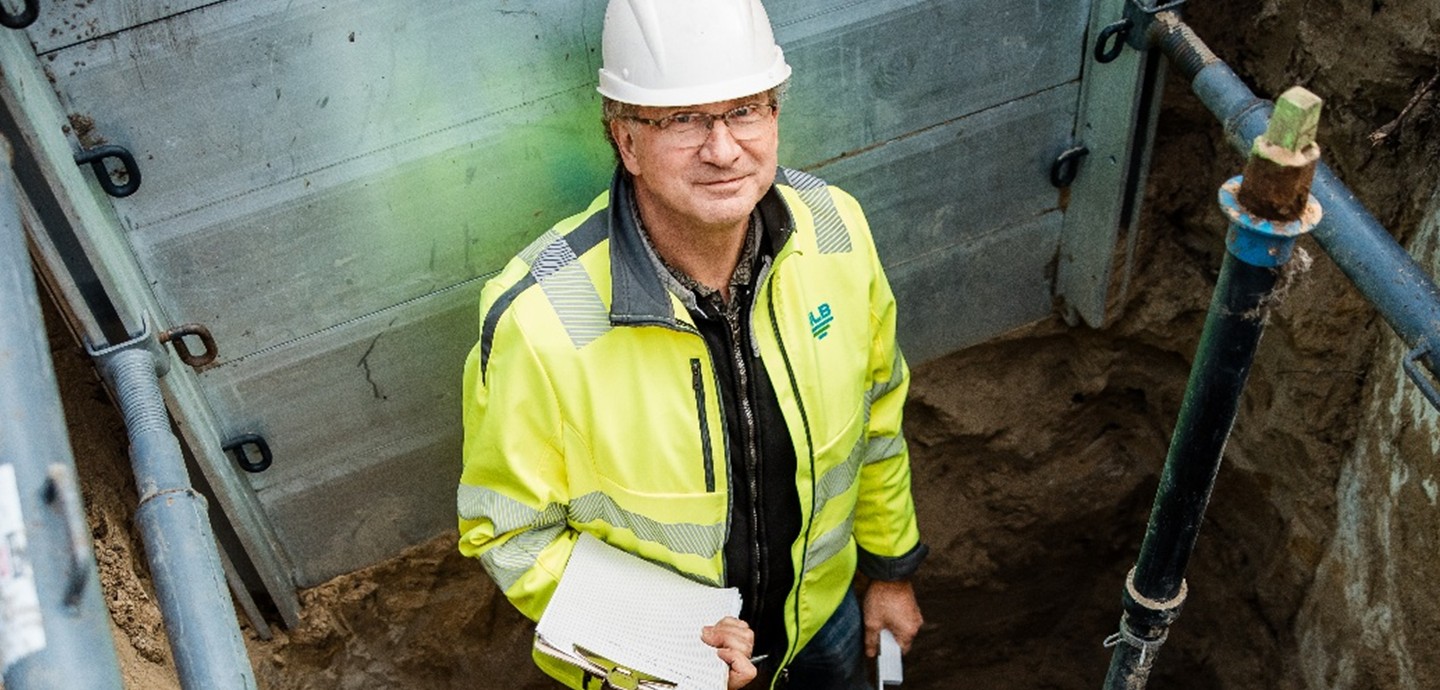 Mann mit Helm und Klemmbrett steht in Grube und lächelt in Kamera